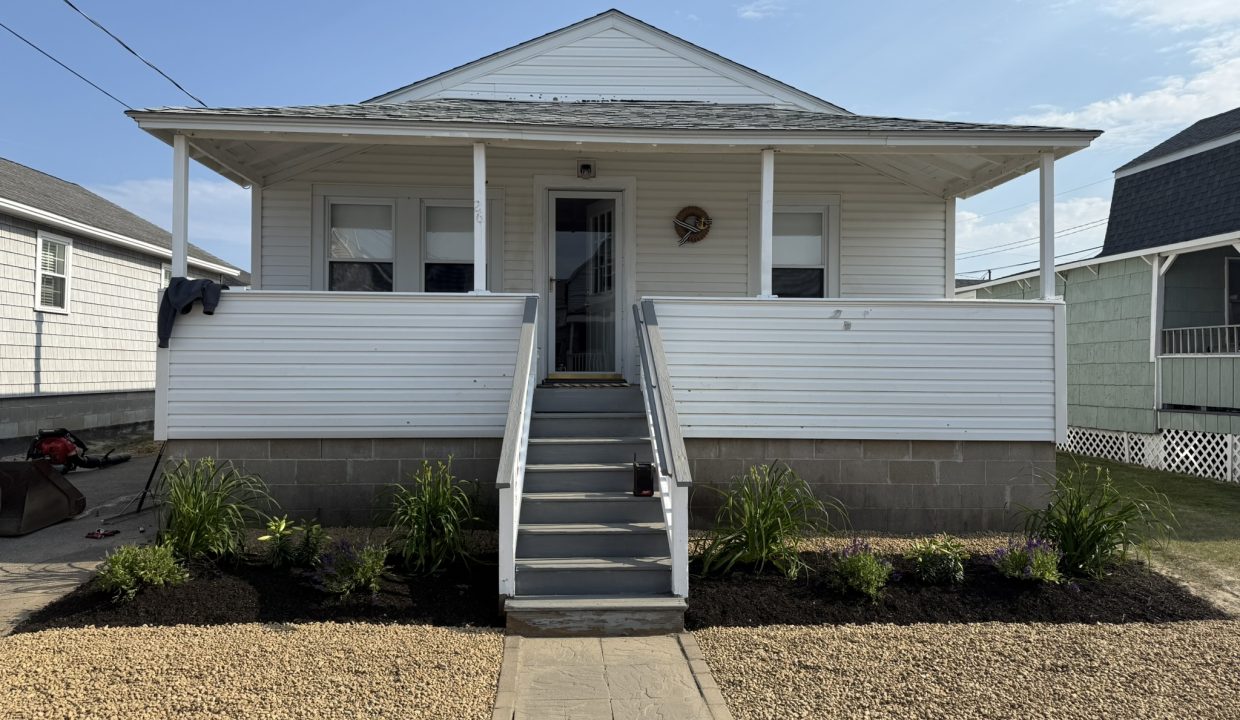 A small white house with a front porch, gray steps, white railing, and a newly landscaped yard with green plants and fresh mulch.