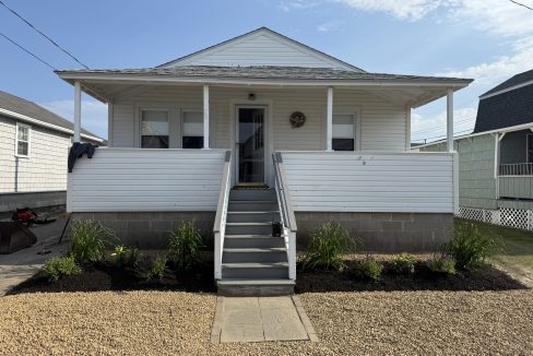 A small white house with a front porch, gray steps, white railing, and a newly landscaped yard with green plants and fresh mulch.