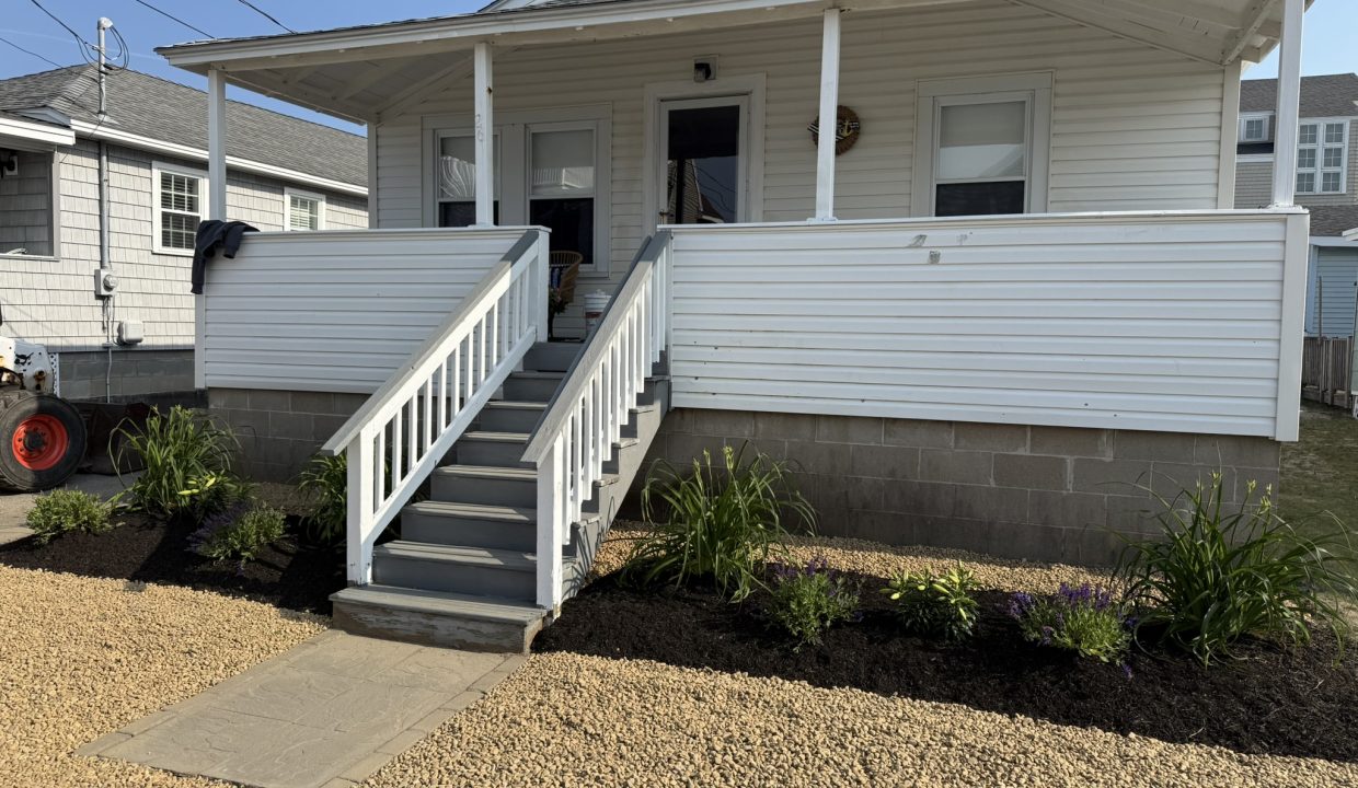A white house with a front porch and stairs, landscaped flower beds on either side, and a gravel walkway and yard leading up to the entrance.