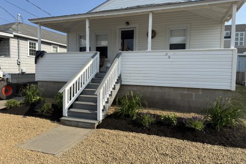 A white house with a front porch and stairs, landscaped flower beds on either side, and a gravel walkway and yard leading up to the entrance.