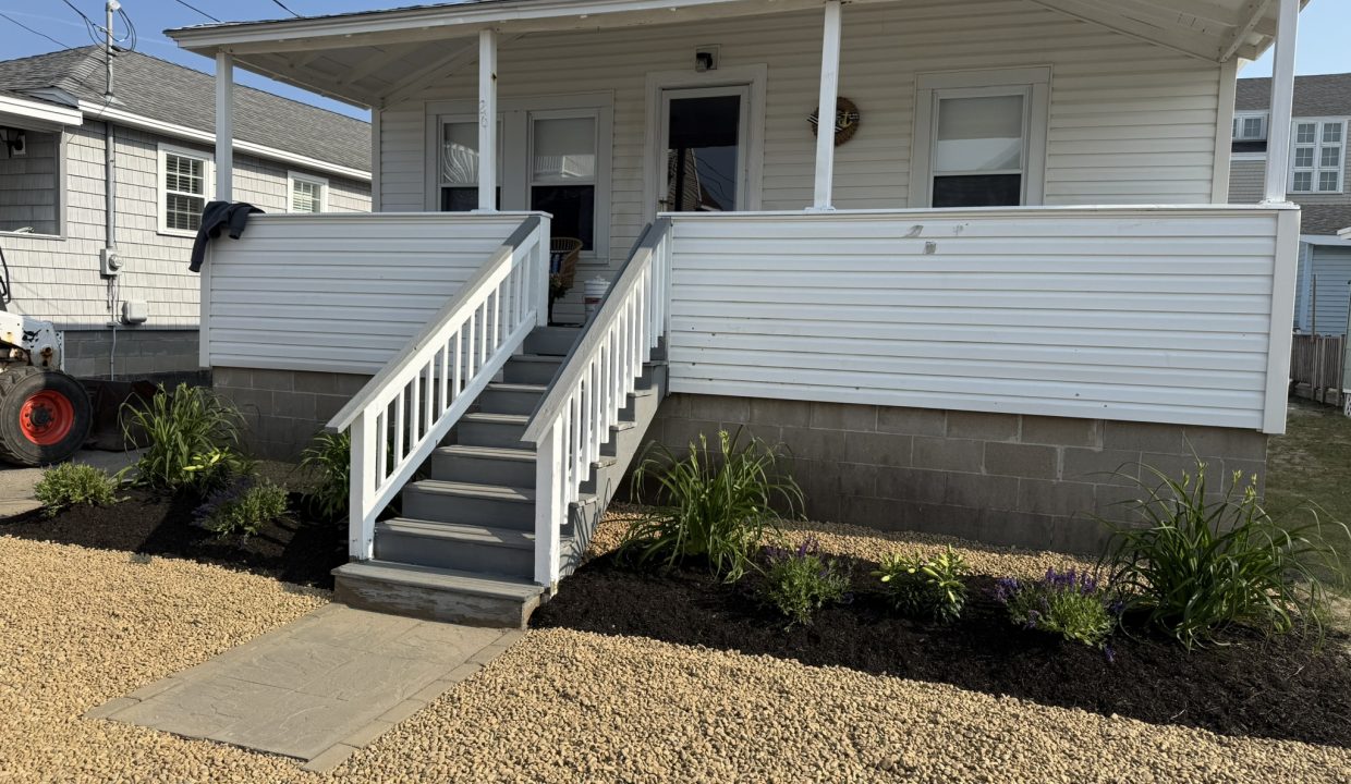 A white house with a front porch, gray stairs, white railing, and landscaped flower beds, set on a gravel yard under clear skies.