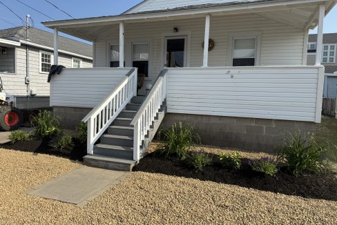 A white house with a front porch, gray stairs, white railing, and landscaped flower beds, set on a gravel yard under clear skies.