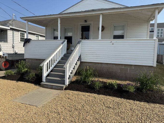 A white house with a front porch, gray stairs, white railing, and landscaped flower beds, set on a gravel yard under clear skies.