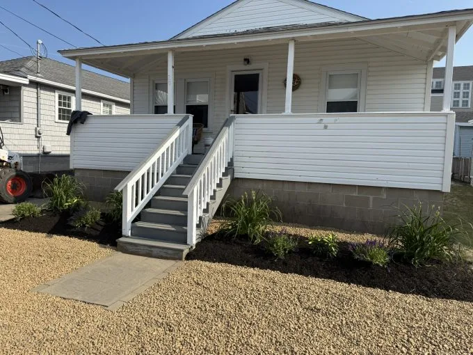 A white house with a front porch, gray stairs, white railing, and landscaped flower beds, set on a gravel yard under clear skies.