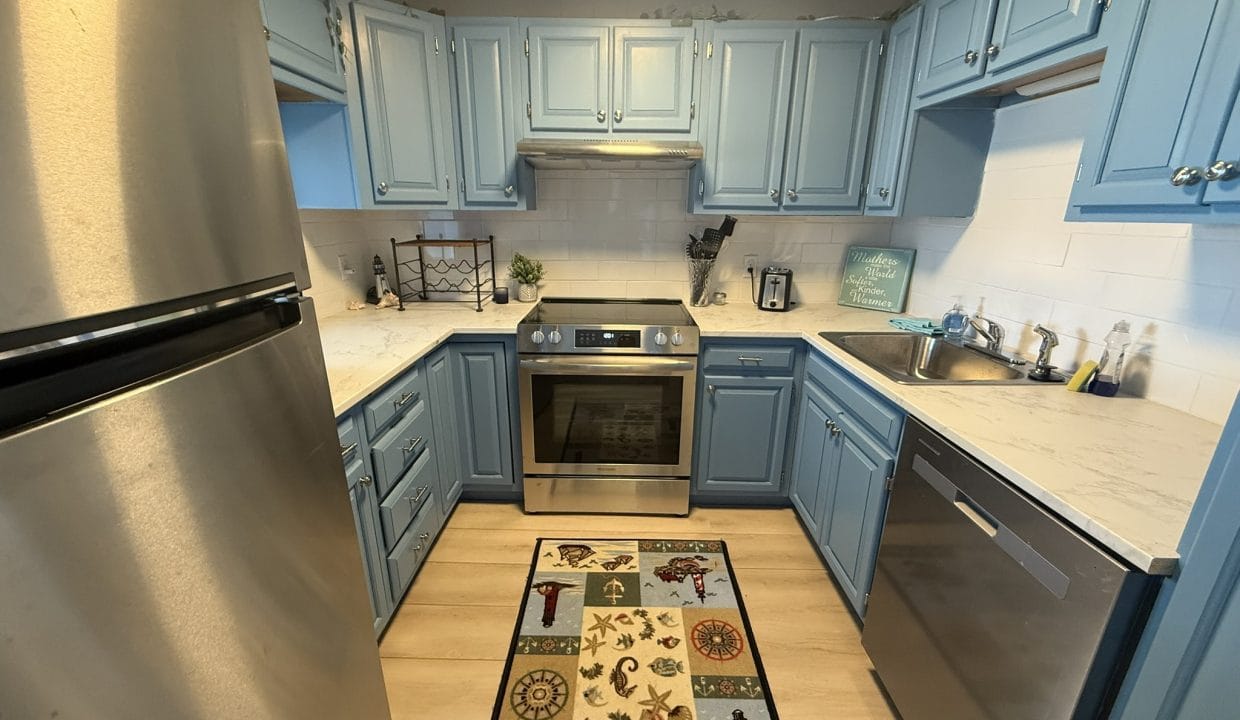 Modern kitchen with light blue cabinets, stainless steel appliances, a patterned rug, and light wood flooring. Countertops are white with subtle grey veining.