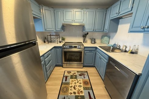 Modern kitchen with light blue cabinets, stainless steel appliances, a patterned rug, and light wood flooring. Countertops are white with subtle grey veining.