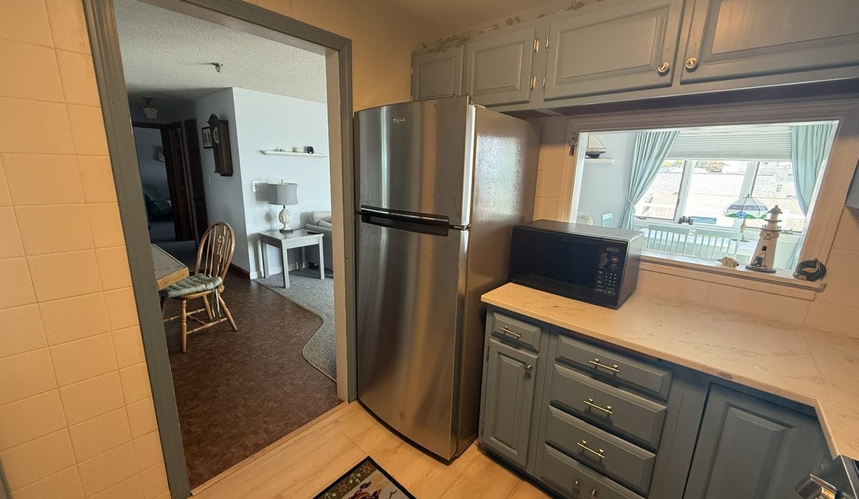 View of a small kitchen with blue cabinets, stainless steel refrigerator, black microwave, and a marble countertop. An adjacent living area is visible through a doorway and pass-through window.
