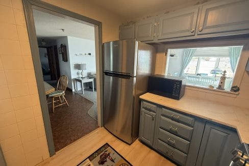 View of a small kitchen with blue cabinets, stainless steel refrigerator, black microwave, and a marble countertop. An adjacent living area is visible through a doorway and pass-through window.