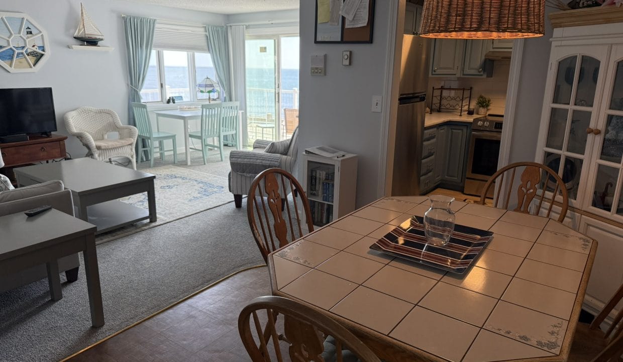 Open-plan living and dining area with wooden table, wicker light fixture, and coastal-themed decor; large windows offer a view of the ocean.