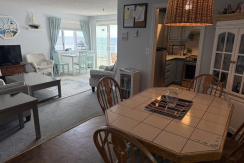 Open-plan living and dining area with wooden table, wicker light fixture, and coastal-themed decor; large windows offer a view of the ocean.