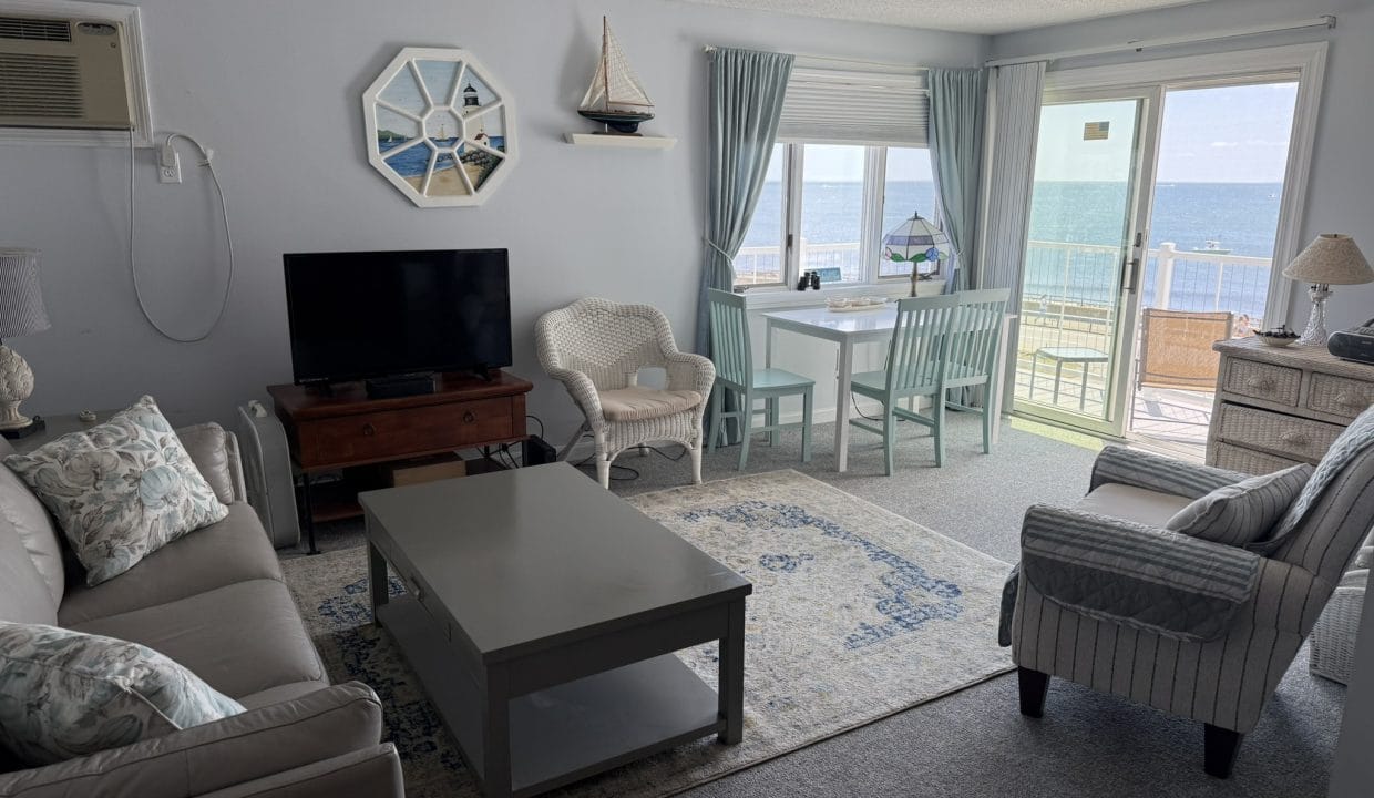 A bright living room with a sofa, armchair, TV, small dining table, and large windows showing an ocean view. Light blue and white decor with nautical accents.