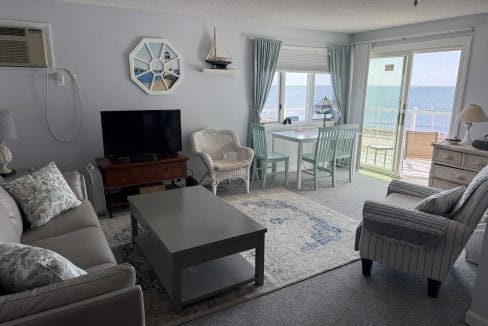 A bright living room with a sofa, armchair, TV, small dining table, and large windows showing an ocean view. Light blue and white decor with nautical accents.