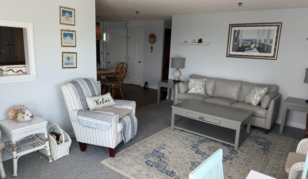A living room with a striped armchair, light gray sofa, coffee table, rug, side tables, wall art, and coastal-themed decor. A dining area is visible in the background.