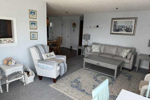 A living room with a striped armchair, light gray sofa, coffee table, rug, side tables, wall art, and coastal-themed decor. A dining area is visible in the background.