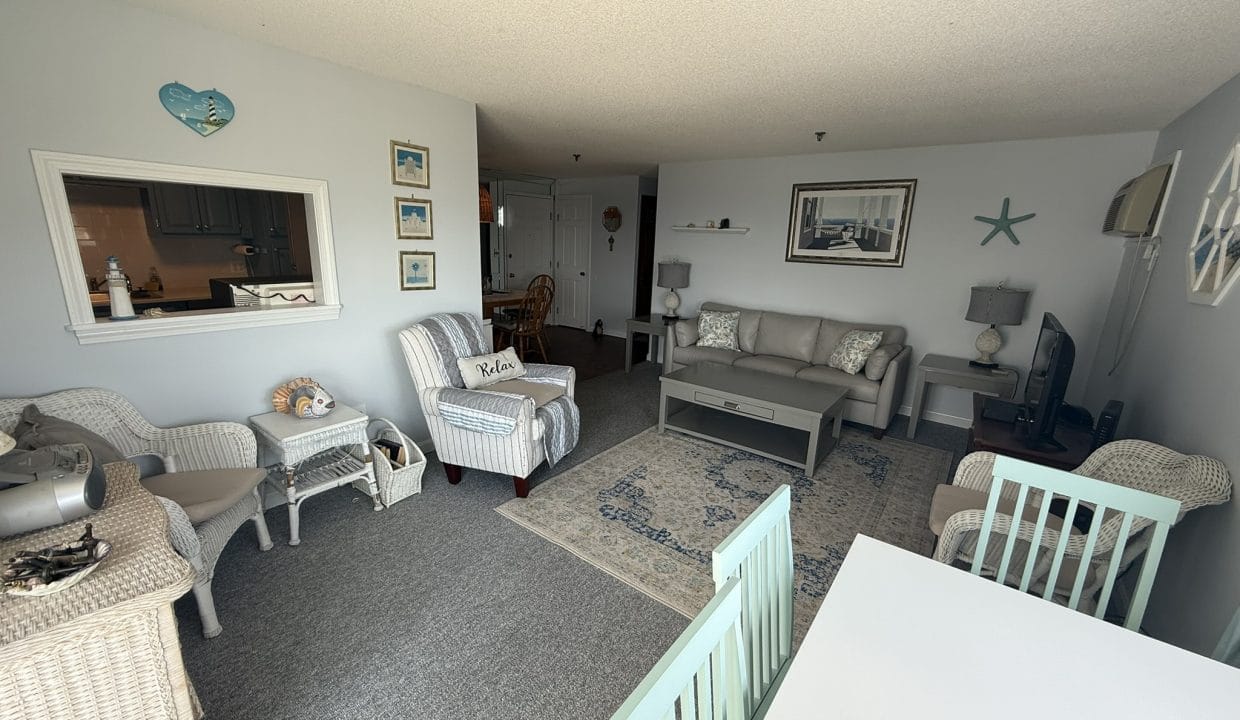 A cozy living room with wicker and upholstered furniture, nautical decor, a TV, and a dining area with mint-green chairs, all in a light, airy setting.