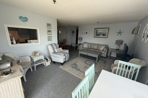 A cozy living room with wicker and upholstered furniture, nautical decor, a TV, and a dining area with mint-green chairs, all in a light, airy setting.