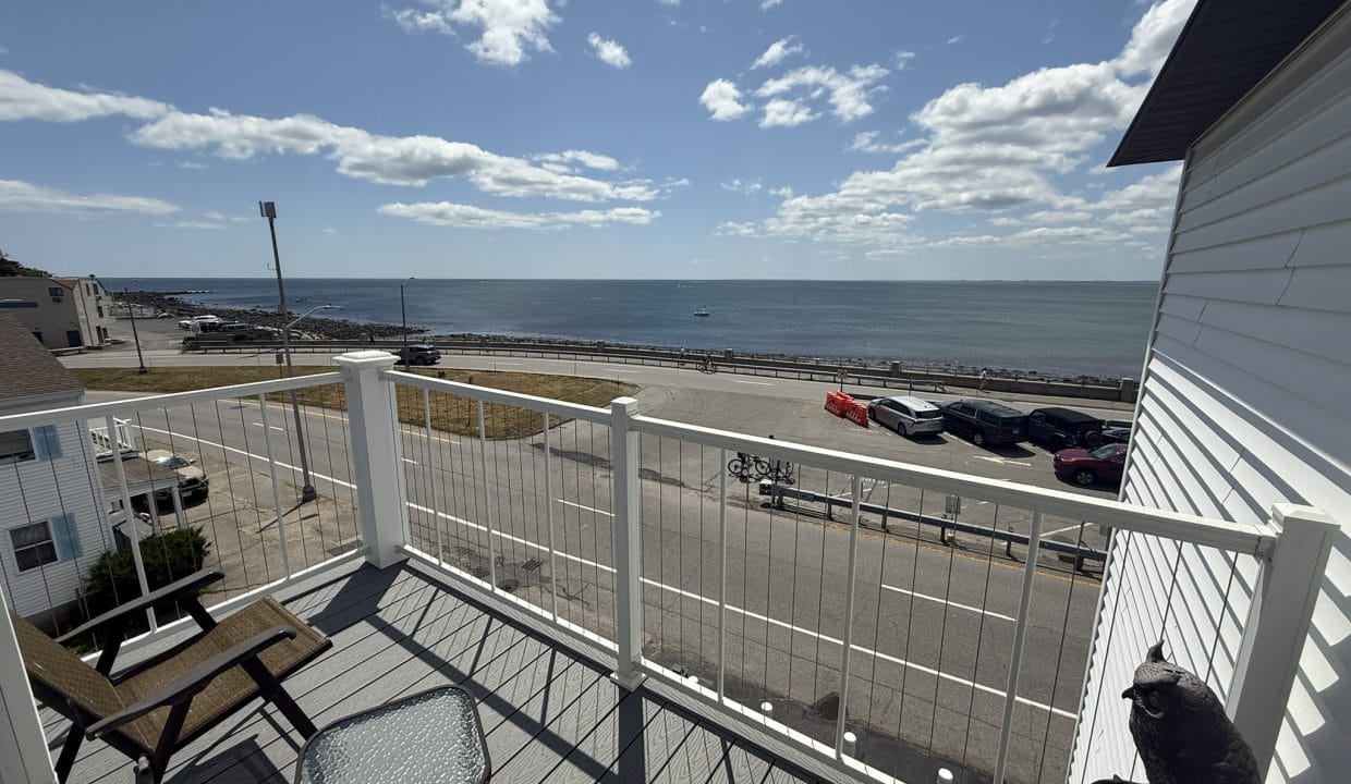 View from a balcony with chairs and a table, overlooking a road, parked cars, and the ocean under a partly cloudy sky.