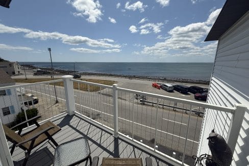 View from a balcony with chairs and a table, overlooking a road, parked cars, and the ocean under a partly cloudy sky.