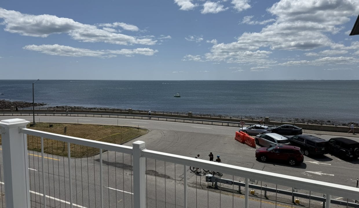View from a balcony overlooking a road, parked cars, grassy area, and the ocean under a partly cloudy sky. A boat is visible in the distance on the water.