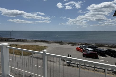 View from a balcony overlooking a road, parked cars, grassy area, and the ocean under a partly cloudy sky. A boat is visible in the distance on the water.