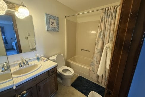 A small bathroom with a sink, mirror, toilet, bathtub with shower curtain, towel, and blue floor mat.