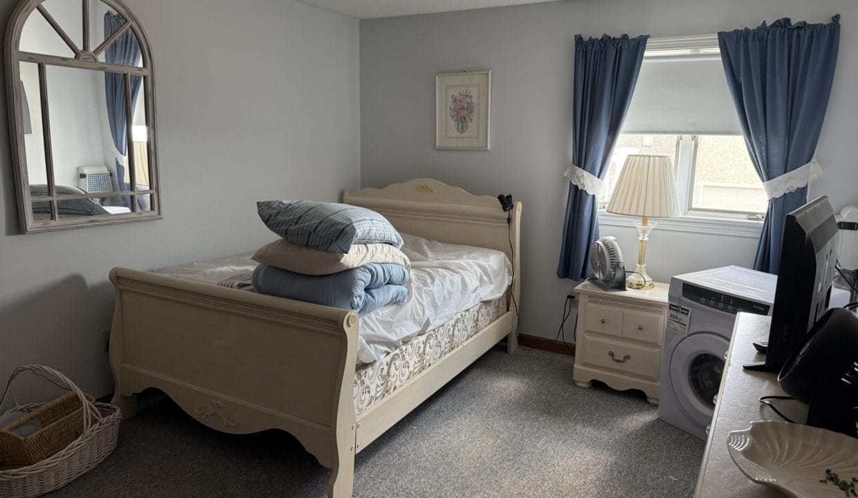 A neatly made bed with folded linens and pillows sits in a small bedroom with blue curtains, a mirror, dresser, laundry basket, and a washing machine near the window.
