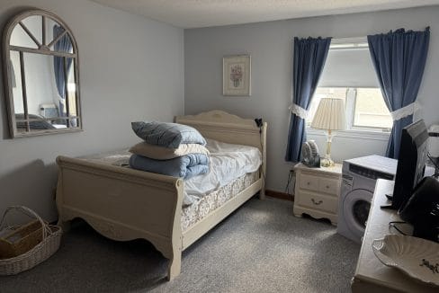 A neatly made bed with folded linens and pillows sits in a small bedroom with blue curtains, a mirror, dresser, laundry basket, and a washing machine near the window.