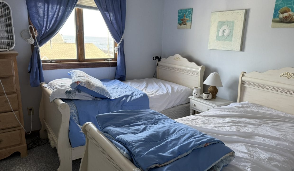 A bedroom with two twin beds, each with blue and white bedding, a window with blue curtains, and light-colored furniture including a nightstand and dresser.