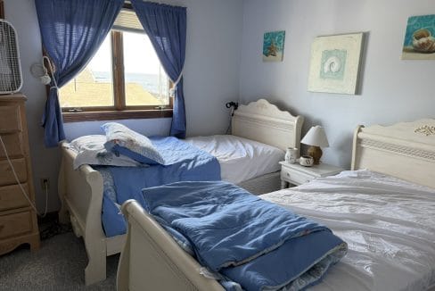 A bedroom with two twin beds, each with blue and white bedding, a window with blue curtains, and light-colored furniture including a nightstand and dresser.
