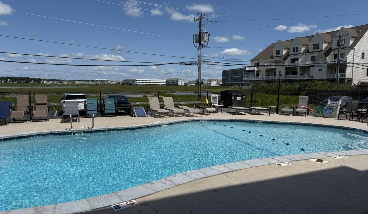 Outdoor swimming pool surrounded by lounge chairs, with parked cars, grassy field, and residential buildings in the background under a sunny sky.