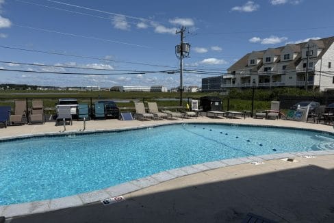 Outdoor swimming pool surrounded by lounge chairs, with parked cars, grassy field, and residential buildings in the background under a sunny sky.