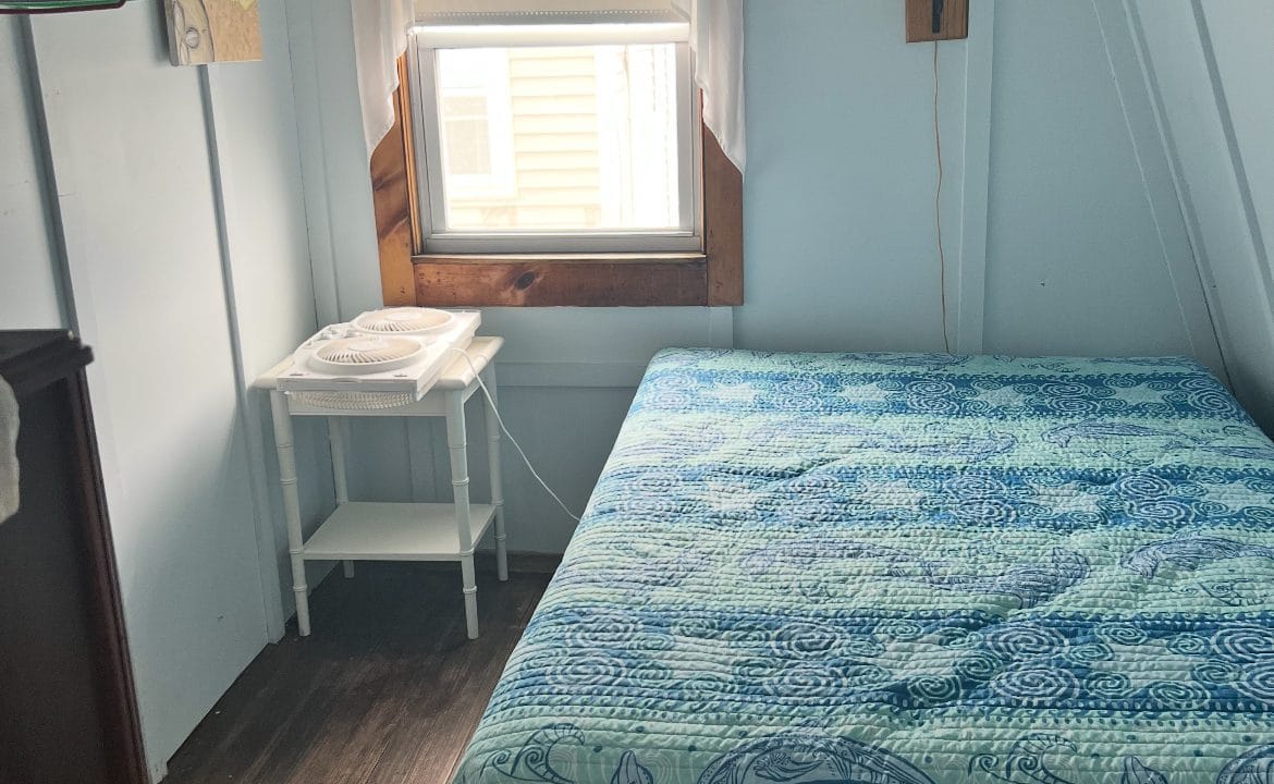 Small bedroom with a bed covered in a blue whale-patterned quilt, a white side table with a fan, window with curtains, and a few hangers on a wall rack.