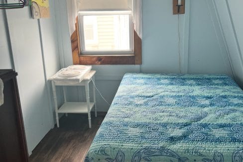 Small bedroom with a bed covered in a blue whale-patterned quilt, a white side table with a fan, window with curtains, and a few hangers on a wall rack.