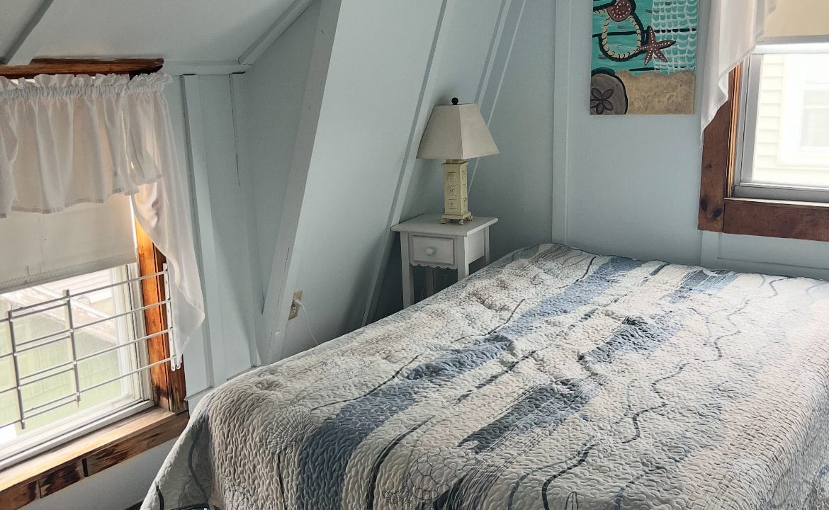 A small bedroom with slanted white walls, a double bed with a patterned quilt, a nightstand with a lamp, two windows, and a wall art piece featuring a bird.