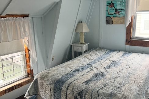 A small bedroom with slanted white walls, a double bed with a patterned quilt, a nightstand with a lamp, two windows, and a wall art piece featuring a bird.