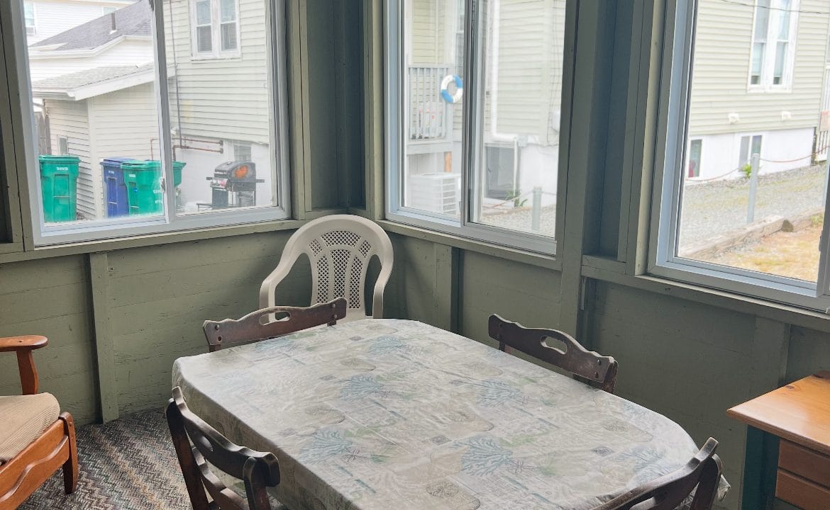 A sunroom with four wooden chairs around a rectangular table covered with a patterned tablecloth, a plastic chair, a ceiling fan, and large windows on three walls.