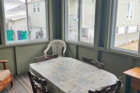 A sunroom with four wooden chairs around a rectangular table covered with a patterned tablecloth, a plastic chair, a ceiling fan, and large windows on three walls.
