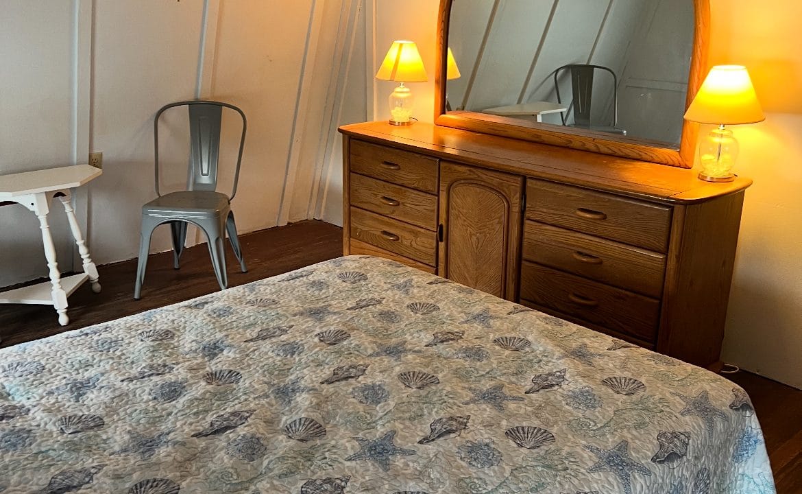 Bedroom with a wooden dresser, round mirror, two lit lamps, metal chair, side table, and bed with a seashell-patterned quilt. White walls and dark wood floor are visible.