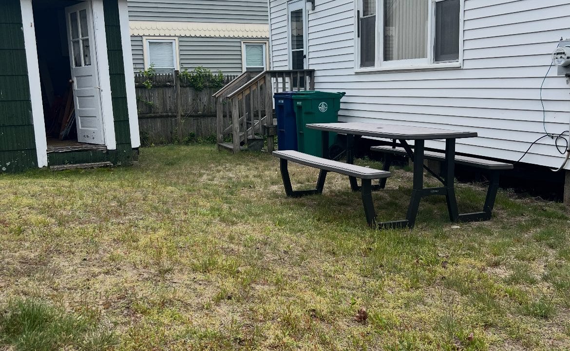 A backyard with patchy grass, a green trash bin, picnic table, and the side of a white house; a small green shed and another building are visible in the background.