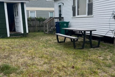 A backyard with patchy grass, a green trash bin, picnic table, and the side of a white house; a small green shed and another building are visible in the background.