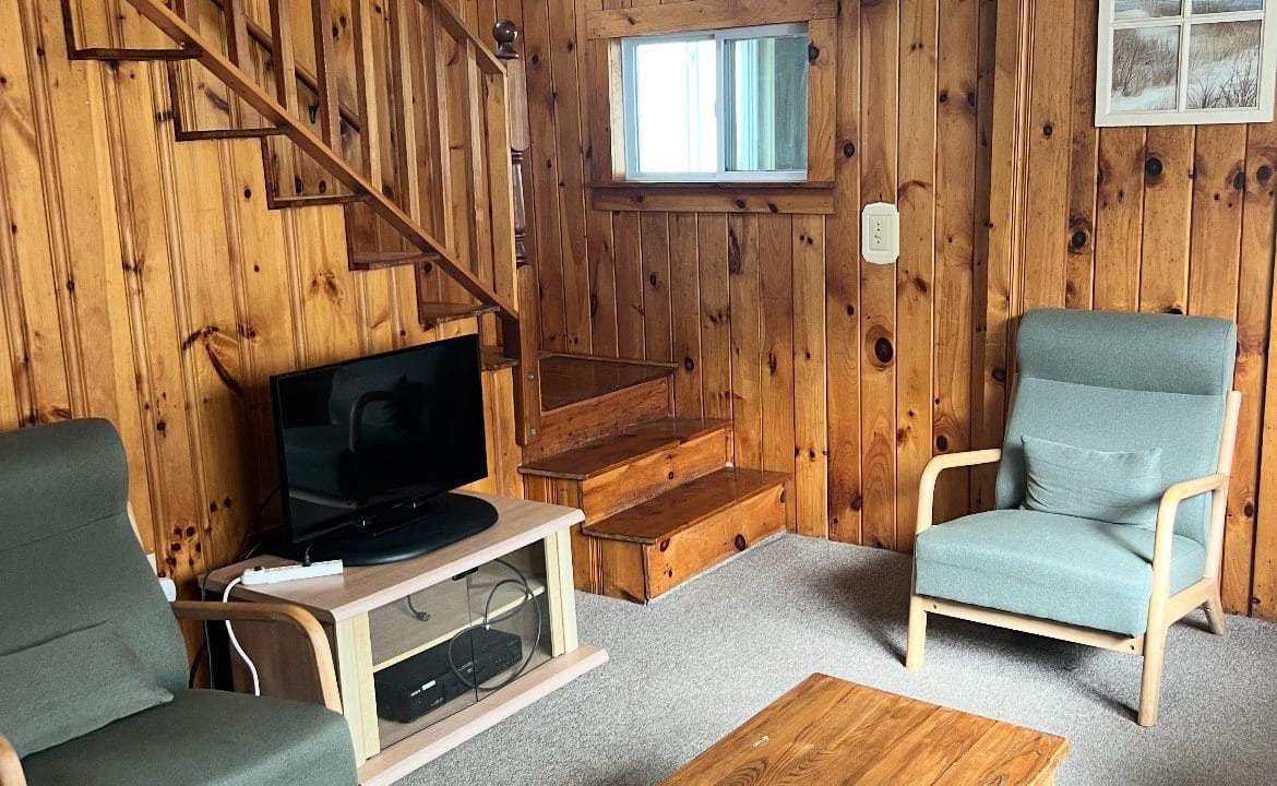 A cozy living room with wood-paneled walls, two armchairs, a wooden coffee table, a TV, and a staircase. A sign reading 
