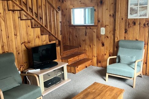A cozy living room with wood-paneled walls, two armchairs, a wooden coffee table, a TV, and a staircase. A sign reading 