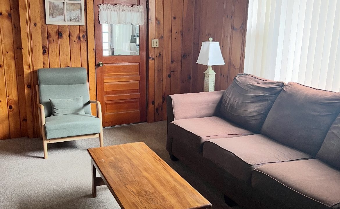 A living room with wood-paneled walls, a brown sofa, a green armchair, a wooden coffee table, a lamp, framed art, and a door with a window and curtains.