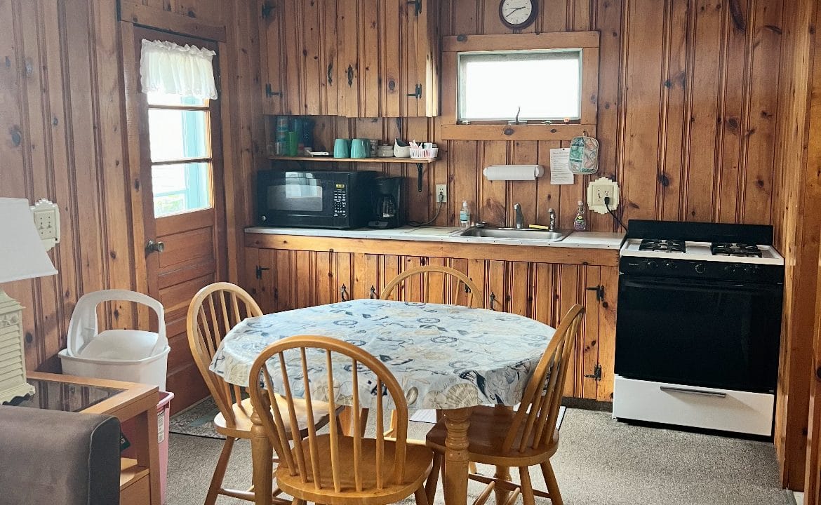 A small kitchen with wood-paneled walls, a table with four chairs, a microwave, stove, sink, and ceiling fan, lit by two windows.
