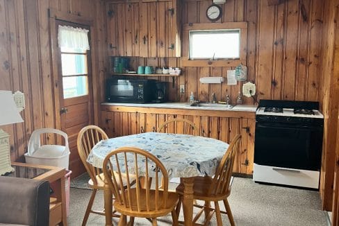 A small kitchen with wood-paneled walls, a table with four chairs, a microwave, stove, sink, and ceiling fan, lit by two windows.