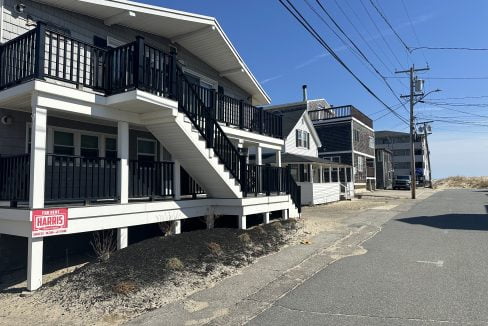 A quiet street lined with two-story residential buildings featuring balconies, with a real estate sign indicating a property for sale.