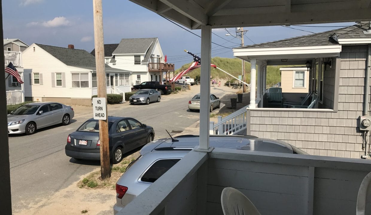 View from a porch overlooking a quiet street with parked cars, houses, and a sand dune path in the background under a partly cloudy sky.