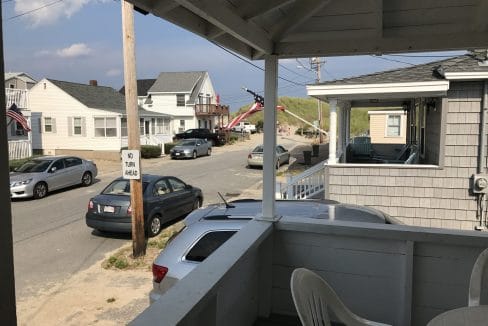 View from a porch overlooking a quiet street with parked cars, houses, and a sand dune path in the background under a partly cloudy sky.