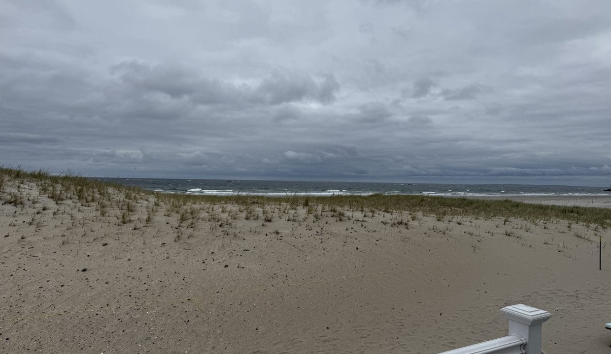A cloudy sky over a sandy beach with sparse grass and ocean waves in the distance; a white railing is visible in the foreground.
