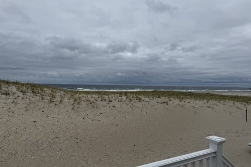 A cloudy sky over a sandy beach with sparse grass and ocean waves in the distance; a white railing is visible in the foreground.